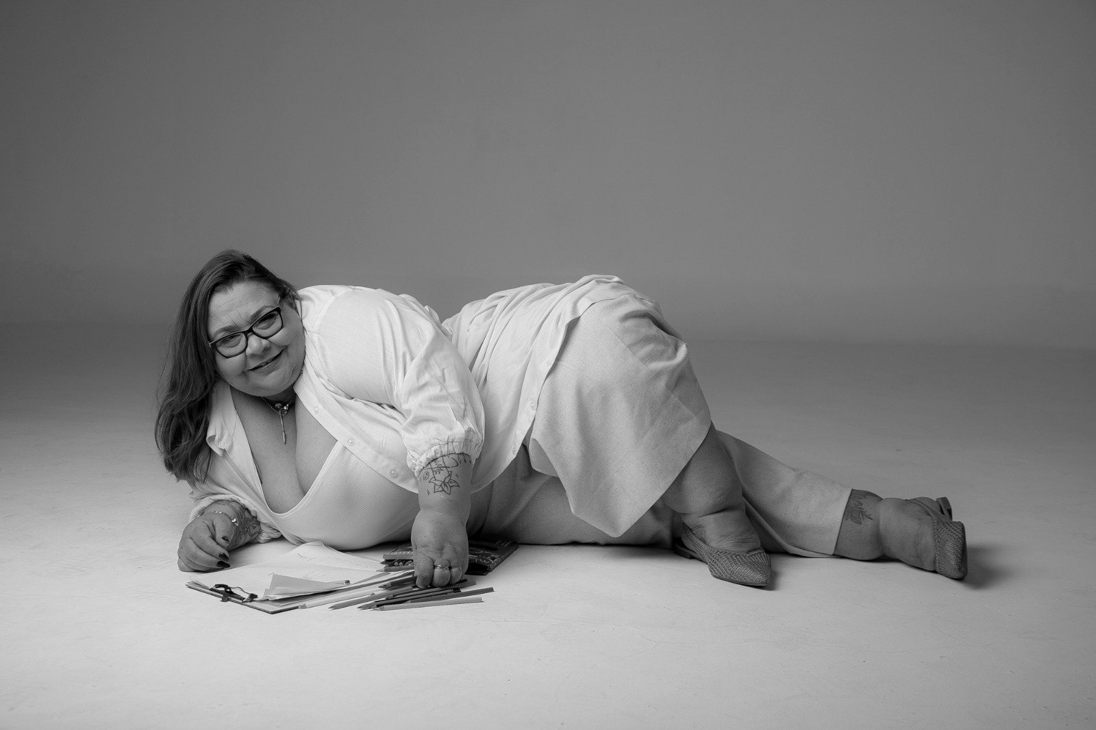 Mulher plus size deitada no chão de estúdio, sorrindo, cercada por papéis e lápis de cor, em preto e branco.