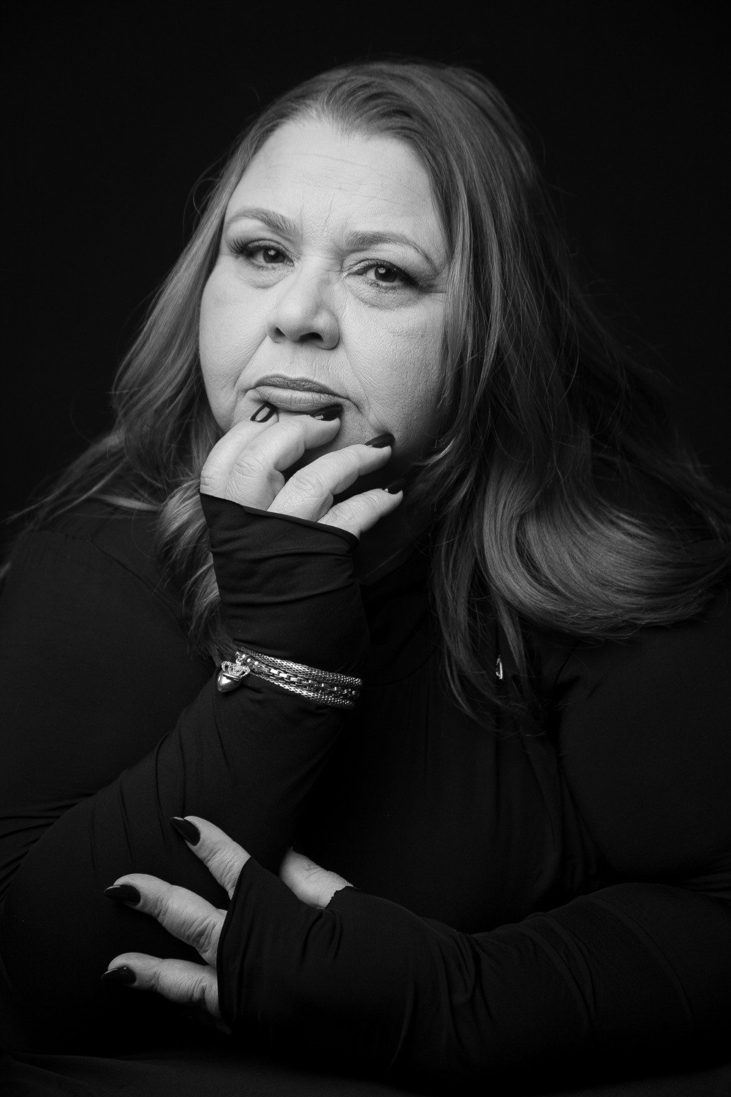 Retrato em preto e branco de mulher plus size, mão apoiada no queixo, olhar direto e presença marcante contra fundo escuro.