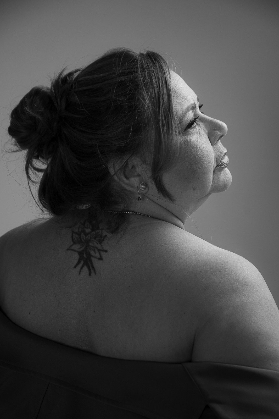 Retrato em preto e branco de mulher plus size, de costas e perfil, tatuagem no ombro, olhar pensativo voltado para longe da câmera.